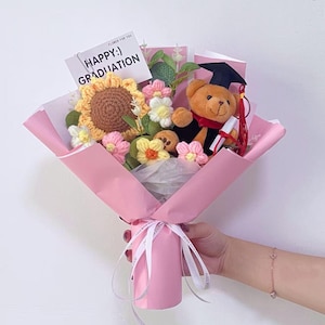 Può includere: Un bouquet di laurea avvolto in carta rosa, legato con un nastro bianco. Il bouquet include un girasole all'uncinetto, vari fiori all'uncinetto e un orsacchiotto con cappello e toga di laurea. Una carta dice "HAPPY :) GRADUATION".
