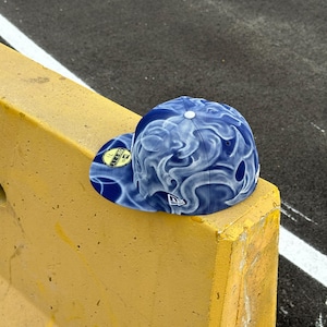 Puede incluir: Una gorra de béisbol ajustada de color azul y blanco con un patrón de humo arremolinado en la corona y la visera. La gorra incluye un botón blanco en la parte superior, un pequeño logotipo blanco bordado en el lateral y una pegatina circular en la visera plana.