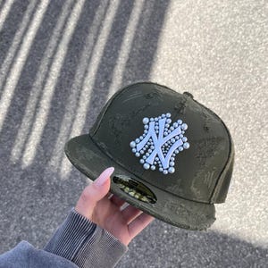 Könnte beinhalten: Eine olivgrüne Baseballkappe mit abgenutzter, ausgefranster Stoffoberfläche. Auf der Vorderseite befindet sich ein weißes New York Yankees Logo, das mit kleinen, runden weißen Perlen verziert ist. Ein runder Aufkleber ist auf dem flachen Schirm.