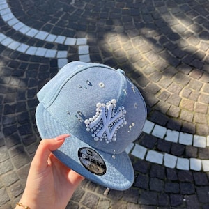 Puede incluir: Una gorra de béisbol de mezclilla azul claro con un logotipo blanco de los New York Yankees adornado con pequeñas perlas blancas y pedrería transparente. La gorra incluye una pegatina 9FIFTY negra y plateada en la visera plana y pedrería dispersa por el frente.