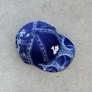 May include: A deep blue baseball cap featuring a white embroidered LA logo on the front. The fabric displays a white airbrushed pattern of chain links, starbursts, and circular shapes across the crown and the flat brim of the hat.