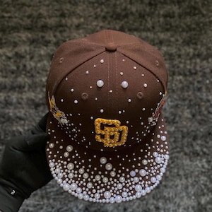 Könnte beinhalten: Eine braune Baseballkappe mit einem gold gestickten SD-Logo auf der Vorderseite, verziert mit verstreuten weißen Perlen und klaren Strasssteinen auf der Krone und dem Schirm, sowie einer gelben Stern-Grafik an der Seite und einem roten Logo.