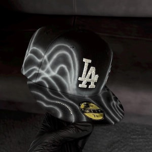 Puede incluir: Una gorra de béisbol negra ajustada con un patrón de ondas aerografiadas en blanco sobre la corona y la visera. El frente muestra un logotipo LA prominente incrustado con pedrería transparente. Una pegatina dorada y negra 59FIFTY está en la visera, talla 7 1/4.