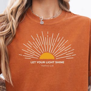 May include: A burnt orange t-shirt featuring the text "LET YOUR LIGHT SHINE" and a sunburst graphic. The shirt is paired with denim shorts. The design includes the Bible verse Matthew 5:16. The t-shirt is made of a soft material.