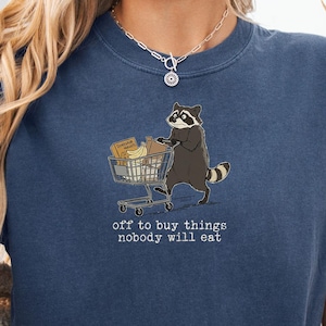May include: Denim blue t-shirt featuring a raccoon pushing a shopping cart filled with groceries. The text "off to buy things nobody will eat" is below the graphic. The shirt is made of a soft material.