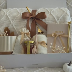 Könnte beinhalten: Geschenkbox mit einer weißen Tasse, einem kleinen Beutel, einem Honigglas, einem Notizblock und einem Stift. Die Box ist mit einem braunen Band und einem goldenen Löffel verziert. Die Tasse hat vertikale Linien und einen braunen Boden.