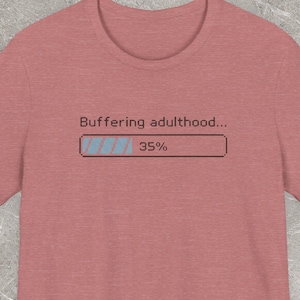 Może przedstawiać: Krótki rękaw, koszulka w kolorze przydymionego różu z napisem "Buffering adulthood..." nad grafiką paska ładowania, która pokazuje 35%. Koszulka wykonana jest z miękkiego materiału i ma okrągły dekolt.