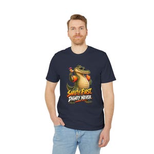 Peut inclure: Un t-shirt bleu marine à manches courtes avec un motif représentant un alligator vert portant des brassards orange et tenant une boisson tropicale avec un petit parasol. Le texte sous l'alligator indique "SAFETY FIRST. DIGNITY NEVER."