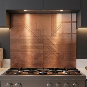 May include: A copper-colored backsplash with a geometric design above a stainless steel gas stovetop. The kitchen features dark cabinets, a white countertop, and wooden cutting boards.