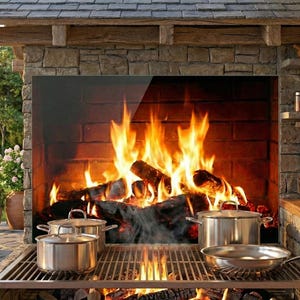 May include: Outdoor kitchen with a stone fireplace and a grill. Stainless steel pots and a frying pan sit on the grill. A fire burns in the fireplace. A wooden table and benches are in the foreground.