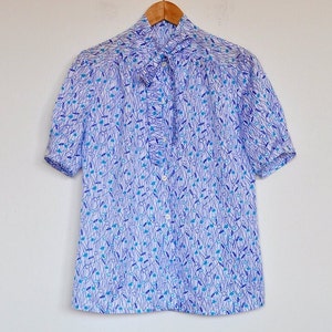 Könnte beinhalten: Ein kurzärmeliges Hemd mit blauem und weißem Blumenmuster. Das Hemd hat eine Rüschendetail auf der Vorderseite und hängt an einem Holzbügel. Das Muster des Hemdes weist blaue und türkisfarbene Akzente auf.