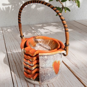 May include: A set of orange and white ceramic ashtrays with a woven handle. The ashtrays are stacked on top of each other and have a heart design.