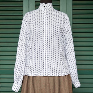 Op de afbeelding: Een wit overhemd met lange mouwen, een hoge kraag en een geometrisch patroon in blauw en wit. Het overhemd is op een mannequin en voor een groene luiken achtergrond.