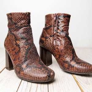 Könnte beinhalten: Ein Paar braune Stiefeletten aus Schlangenleder mit Blockabsatz. Die Stiefeletten haben einen Reißverschluss an der Innenseite jedes Knöchels.