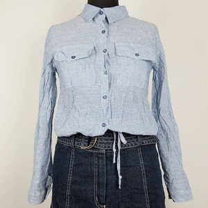 Puede incluir: Una camisa de manga larga de botones a rayas azul claro con dos bolsillos de solapa y un lazo en la cintura. La camisa se lleva sobre unos vaqueros azul oscuro.