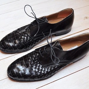 Può includere: Un paio di scarpe eleganti in pelle nera con un motivo intrecciato sulla punta e sulla tomaia. Le scarpe hanno i lacci e sono su uno sfondo di legno chiaro. Uno stile classico.