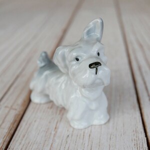 Peut inclure: Petite figurine de chien en céramique blanche, avec des détails gris sur les oreilles et la queue. Le chien a des yeux noirs et un nez noir. La figurine est en position assise.