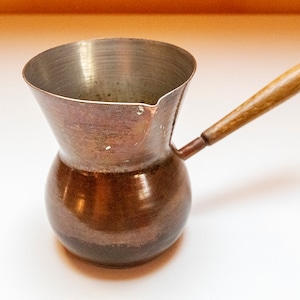 Könnte beinhalten: Eine Kaffeekanne aus Kupfer mit einem Holzgriff. Die Kanne hat einen runden Körper und einen ausgestellten oberen Teil. Der Griff ist an der Seite der Kanne befestigt.