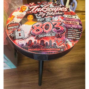 May include: A round side table with a black base and three legs, featuring a vibrant graphic tabletop. The design includes the text Inkscapes By Tekesha, Rep The 803, Columbia, SC, and various icons like a padlock, city skyline, and hearts.