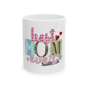 May include: White ceramic mug featuring the text "best MOM ever" in a colourful collage design. The letters are in various patterns, including plaid, leopard print, and floral, with heart and star accents.