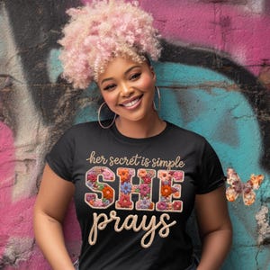 Może przedstawiać: Czarny t-shirt z napisem "her secret is simple SHE prays" w kwiatowym wzorze. Kobieta na zdjęciu uśmiecha się i nosi kolczyki koła. Tło to ściana graffiti z różową i niebieską farbą.