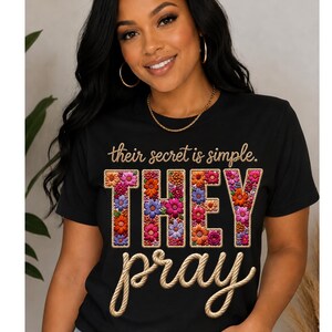 Puede incluir: Una camiseta negra de manga corta con el texto "their secret is simple." en una fuente cursiva color canela. Debajo, la palabra "THEY" aparece en letras grandes rellenas de patrones florales coloridos, seguida de "pray" en una fuente estilo cuerda.