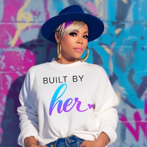 May include: A woman wearing a white sweatshirt with the text "BUILT BY her" in black and gradient blue-purple. She is wearing a blue hat and gold hoop earrings. The background is a colorful graffiti wall.
