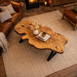 Op de afbeelding: Een unieke salontafel van hout met een natuurlijke rand en een organische vorm. De tafel wordt ondersteund door zwarte metalen poten. Een kleine witte vaas met droogbloemen, een kopje koffie en boeken zijn op de tafel geplaatst. De tafel is ongeveer 122 cm lang.