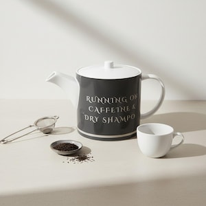Könnte beinhalten: Eine grau-weiße Teekanne mit dem Text "RUNNING ON CAFFEINE & DRY SHAMPOO" steht neben einer weißen Teetasse, einer kleinen Schale mit Teeblättern und einem Teesieb. Die Teekanne hat einen weißen Deckel und Henkel. Das Teeset steht auf einer hellen Oberfläche.