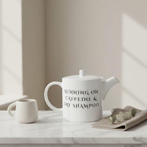 Könnte beinhalten: Weißes Keramik-Teeservice mit Teekanne und Tasse. Die Teekanne hat einen Deckel und einen gebogenen Griff, mit dem schwarzen Text "RUNNING ON CAFFEINE & DRY SHAMPOO". Eine kleine, passende Tasse befindet sich links.