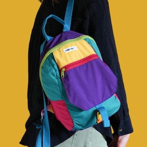 Può includere: Uno zaino multicolore con pannelli in tessuto viola, verde acqua, giallo e rosa. La borsa include una maniglia superiore, spallacci blu, una tasca frontale con cerniera e un'etichetta bianca con il testo nero JOLLYZU sul pannello giallo.