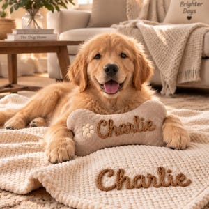 Op de afbeelding: Een golden retriever puppy liggend op een crèmekleurige, getextureerde deken. De puppy rust met zijn poten op een botvormig pluche speeltje, geborduurd met de naam Charlie en een pootafdruk. De deken is ook voorzien van de naam Charlie in bruin garen.