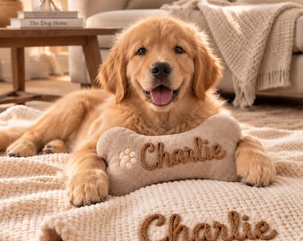 May include: A golden retriever puppy lying on a cream-colored textured blanket, resting its paws on a bone-shaped plush toy. Both the blanket and the toy feature the name Charlie embroidered in brown yarn, with a small paw print on the toy.