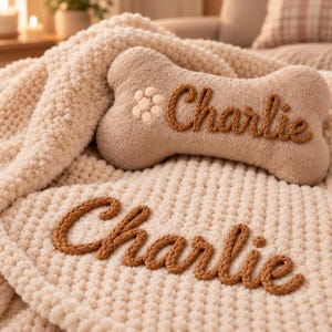 May include: A cream-colored textured blanket and a bone-shaped pillow in a matching shade. Both items feature the name Charlie embroidered in a thick, brown, braided yarn script. The pillow also includes a small, circular, cream-colored flower.