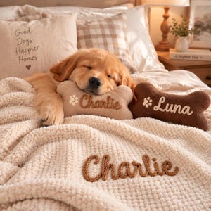 May include: A golden retriever puppy sleeping on a cream-colored textured blanket. Two bone-shaped plush toys are present, one tan embroidered with 'Charlie' and a paw print, and one brown embroidered with 'Luna' and a paw print.