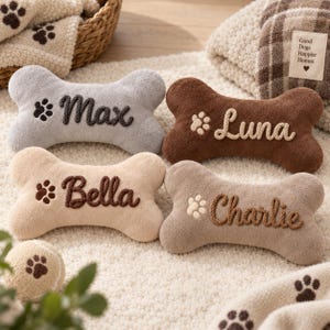 May include: Four plush bone-shaped toys in gray, brown, beige, and tan, each embroidered with a name: Max, Luna, Bella, and Charlie, accompanied by a paw print icon. A small matching ball and a blanket with the text Good Dogs Happier Homes.