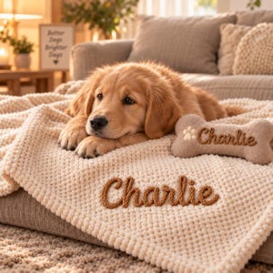 May include: A golden retriever puppy rests on a cream-colored textured blanket featuring the name Charlie embroidered in brown yarn. Beside the puppy lies a bone-shaped toy with a paw print and the name Charlie stitched in brown.