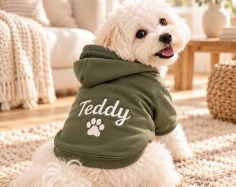 May include: A small white dog wearing an olive green hooded sweatshirt. The back of the hoodie features the name Teddy embroidered in white script lettering above a white paw print graphic. The garment has long sleeves and a ribbed hem.
