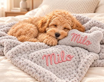 May include: A light gray chunky knit blanket and a matching bone-shaped pillow, both featuring the name Milo embroidered in pink yarn. A golden-brown puppy is sleeping on the soft, textured fabric of the blanket and resting its head on the pillow.