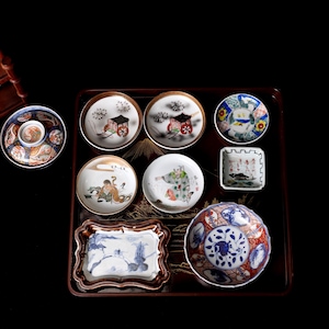 Könnte beinhalten: Eine Sammlung antiker japanischer Porzellanteller und -schalen mit handgemalten Szenen und Designs. Die Teller variieren in Größe und Form, mit komplizierten Details und einer Reihe von Farben, darunter Blau, Rot und Gold. Die Sammlung wird auf einem dunklen Holztablett präsentiert.