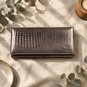 May include: A rectangular metallic bronze wallet with a textured crocodile skin pattern. The wallet features a flap closure and a small silver-toned metal logo plate on the bottom right corner. The surface has a glossy finish and a dark sheen.