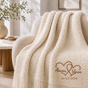 Può includere: Una coperta in maglia color crema con un motivo diagonale testurizzato e frange. La coperta presenta un ricamo di due cuori intrecciati con i nomi Aaron e Venus e la data 10/21/2006 realizzati con filo di colore marrone.
