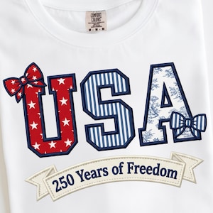 Könnte beinhalten: Weißes T-Shirt mit dem Wort "USA" in roter, weißer und blauer Stickerei. Das "U" hat rote Sterne, das "S" blaue Streifen und das "A" ein blau-weißes Blumenmuster. Der Text "250 Years of Freedom" ist ebenfalls vorhanden.