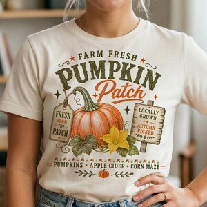 Könnte beinhalten: Ein cremefarbenes T-Shirt mit einem Grafikdruck und dem Text Farm Fresh Pumpkin Patch. Das Design zeigt einen großen orangefarbenen Kürbis, ein Etikett mit der Aufschrift Fresh From The Patch und ein Holzschild mit Locally Grown Autumn Picked.