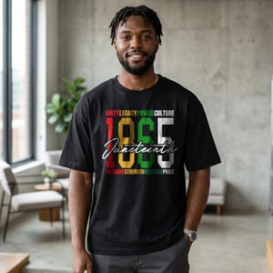 K&ouml;nnte beinhalten: Schwarzes T-Shirt mit dem Text "UNITY LEGACY POWER CULTURE" &uuml;ber den Zahlen "1865" in Rot, Gold, Gr&uuml;n und Wei&szlig;. Das Wort "Juneteenth" ist kursiv unter den Zahlen geschrieben. Die Worte "FREEDOM STRENGTH HERITAGE PRIDE" stehen darunter.