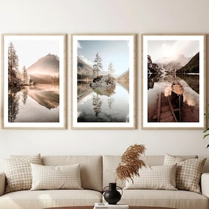 May include: Three framed landscape art prints featuring serene lake scenes with mountain reflections. The prints are in light wood frames and hang above a beige sofa with patterned pillows. A lamp and a potted plant are also in the room.