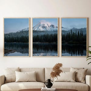 May include: Three framed landscape prints depicting a mountain range with snow-capped peaks reflected in a lake. The artwork features a forest of evergreen trees and a muted color palette of blues and greens. The frames are a light wood color.