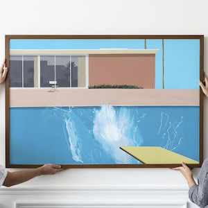 May include: A framed art print showing a mid-century modern home with a swimming pool. The artwork features a blue pool with a splash, a yellow diving board, and a pink and white building. Two people are holding the framed print.