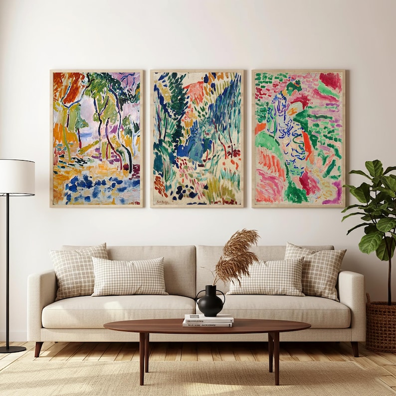 May include: Three framed abstract art prints with vibrant colors hang above a beige sofa. The artwork features a mix of red, green, blue, and yellow hues, depicting a landscape scene. The room is decorated with a floor lamp, a coffee table, and a potted plant.