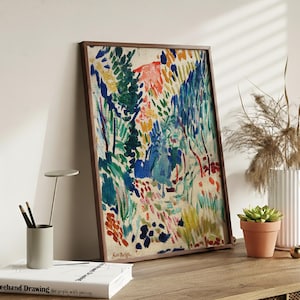 May include: A framed abstract painting with a brown wooden frame. The artwork features a colorful landscape with blue, green, red, and yellow brushstrokes. The painting is leaning against a white wall, with a small succulent in a terracotta pot.